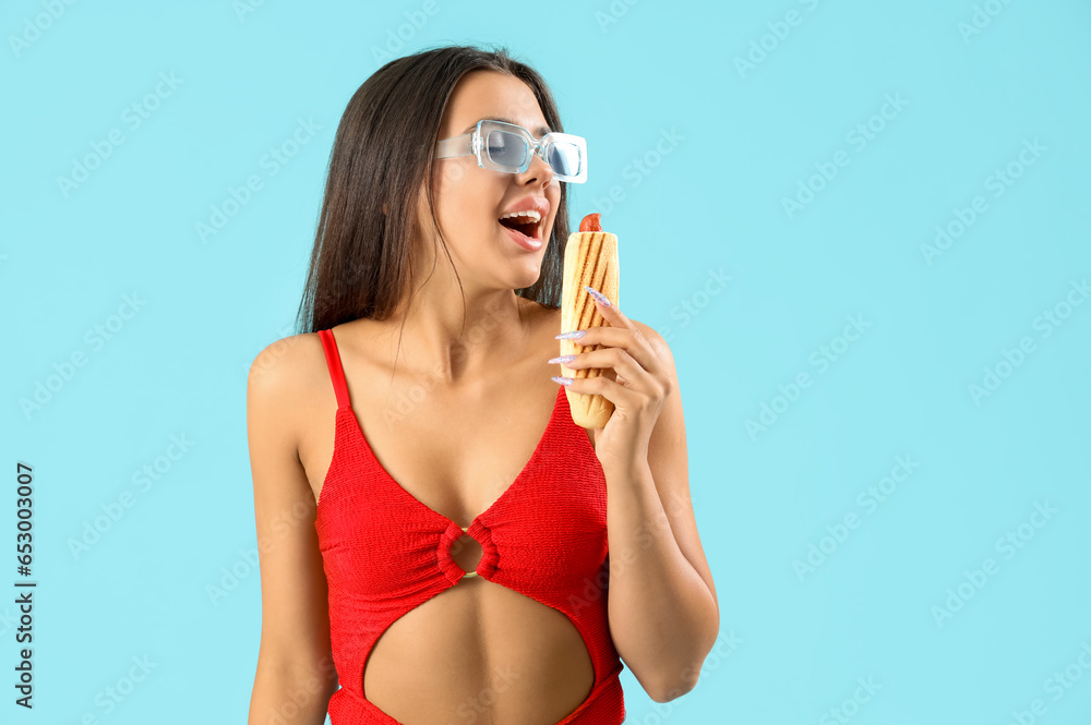 Pretty young woman in swimsuit and with tasty french hot dog on blue background