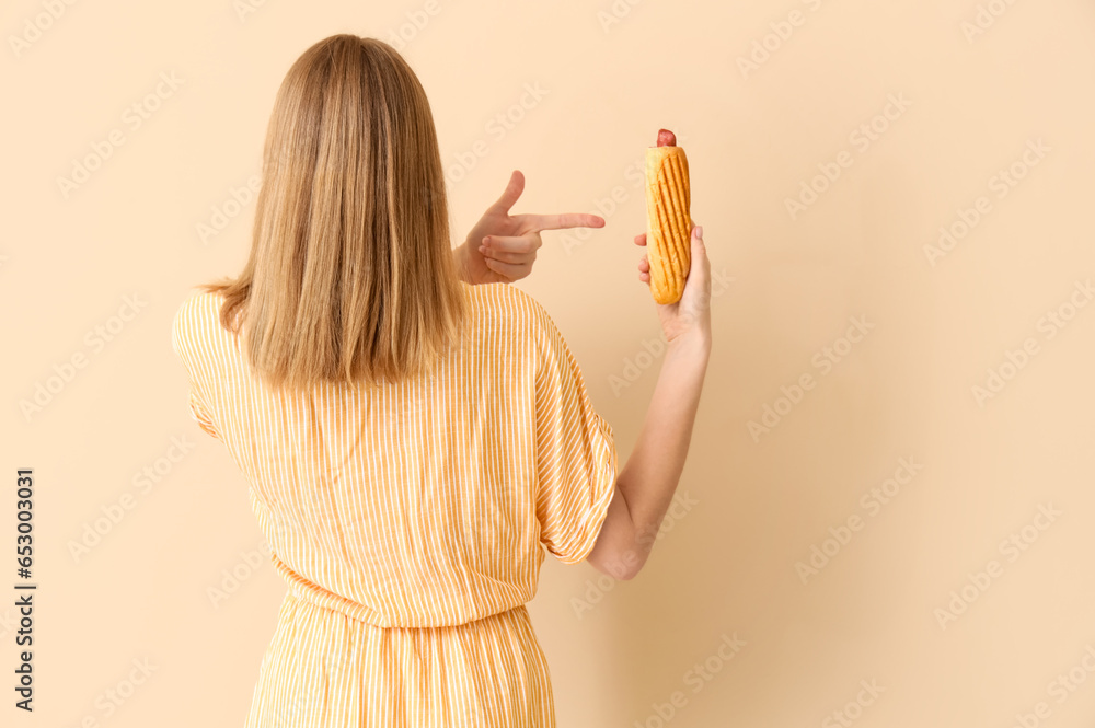 Pretty young woman with tasty french hot dog on beige background, back view