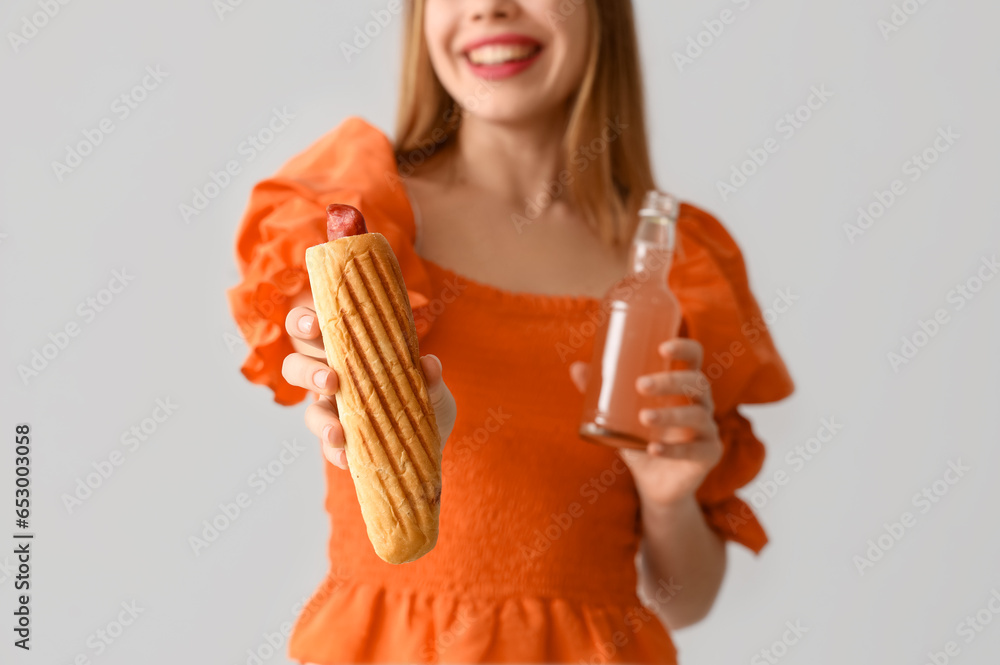 Pretty young woman with tasty hot dog and drink on grey background