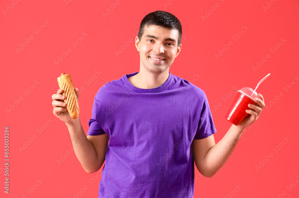 Happy young man with tasty french hot dog and drink on red background