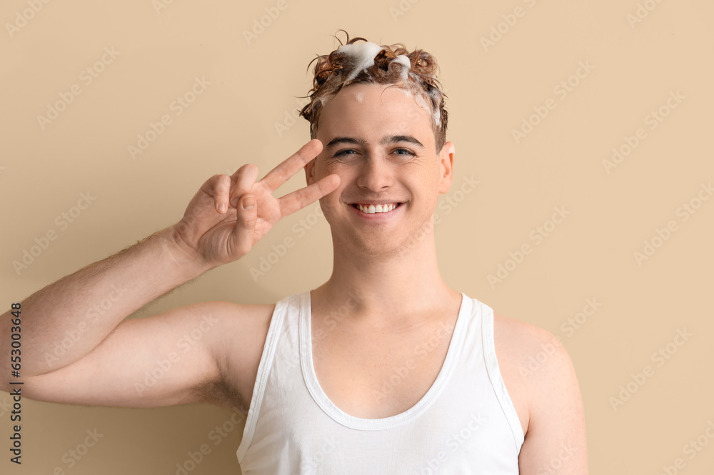 Young man with applied shampoo on his hair showing victory gesture against beige background