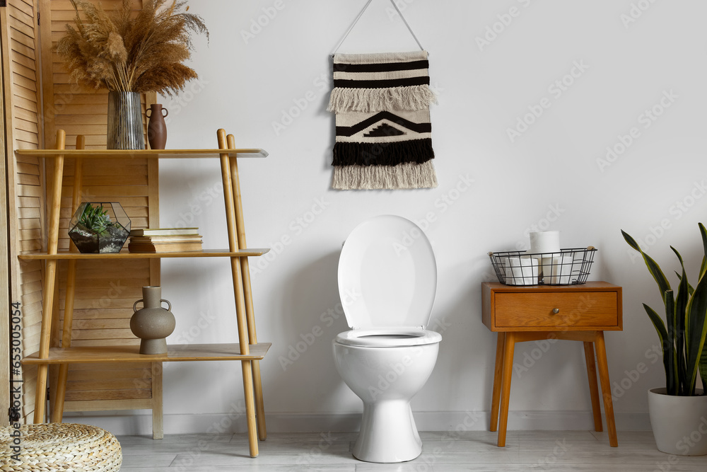 Interior of light restroom with ceramic toilet bowl, paper rolls on table and shelving unit