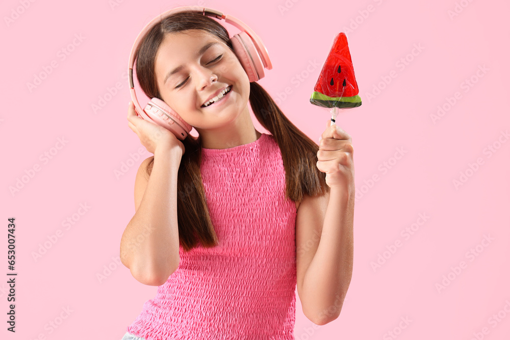 Happy little girl with lollipop in shape of watermelon slice listening music on pink background