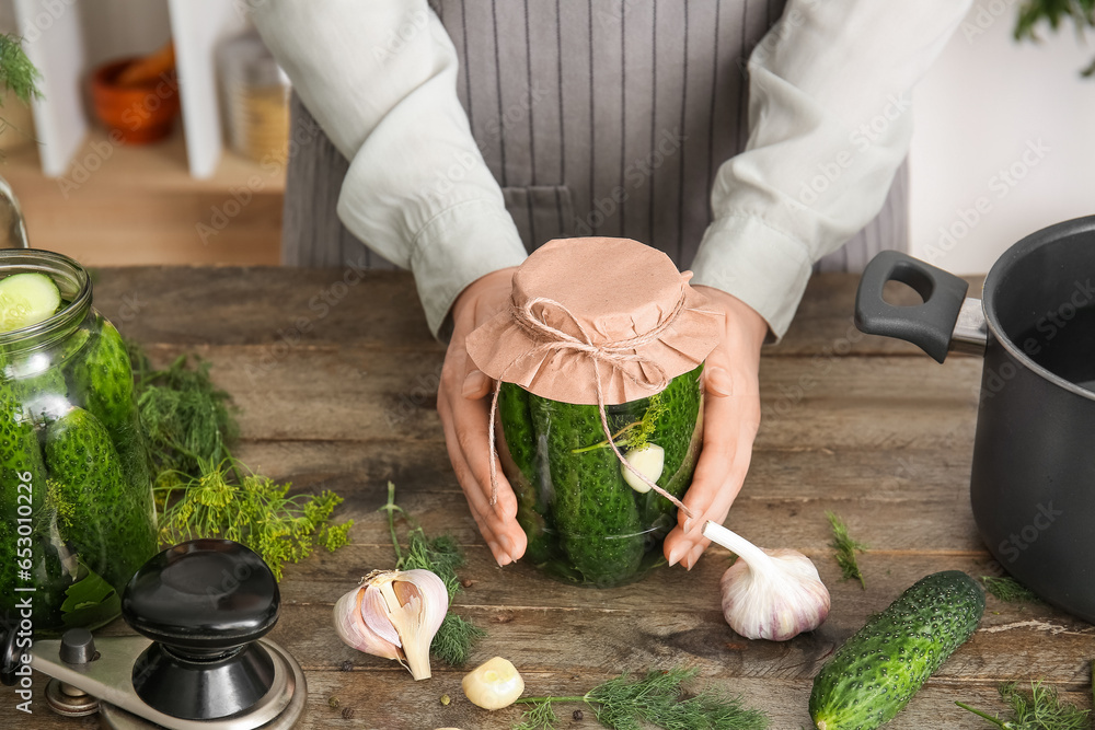 Woman holding jar with canned cucumbers at table in kitchen