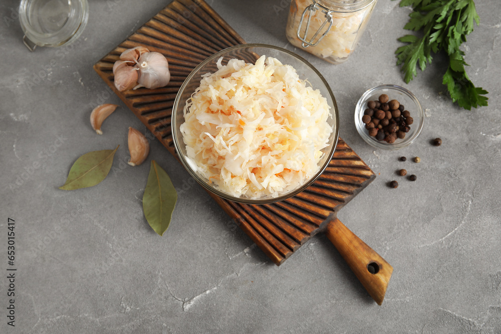 Wooden board with glass bowl of delicious sauerkraut and different spices on grey background