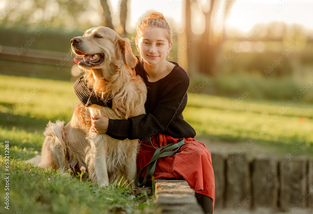 Pretty girl with golden retriever dog sitting at nature and smiling. Beautiful female model teenager hugging purebred pet doggy at autumn park