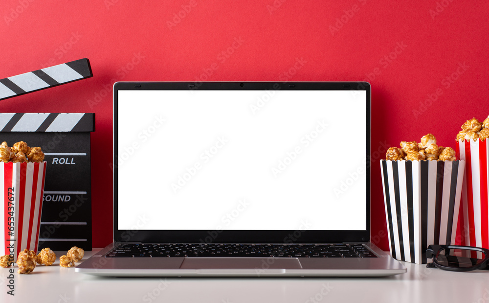 Indoor movie night concept. Side view of a coffee table with a laptop for streaming, boxes with popcorn, 3D glasses and clapperboard with a red wall background with empty screen for text or advert