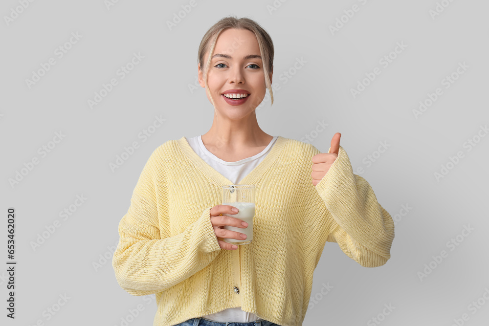 Young woman with glass of milk showing thumb-up on grey background