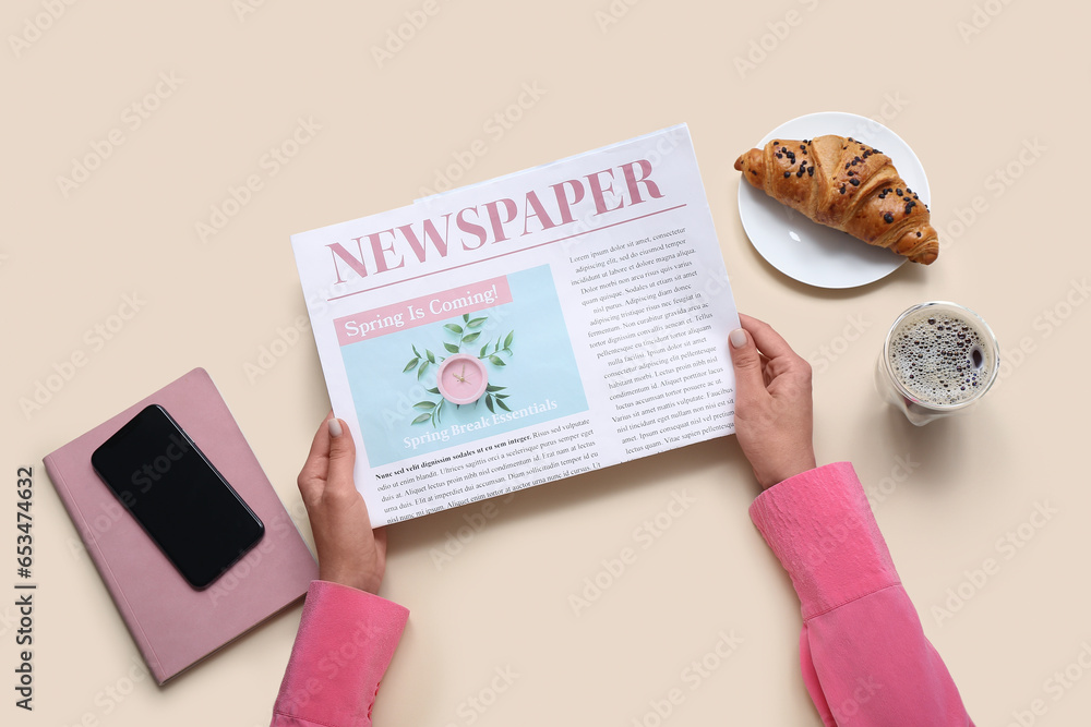 Woman reading newspaper with coffee, croissant and mobile phone on beige background