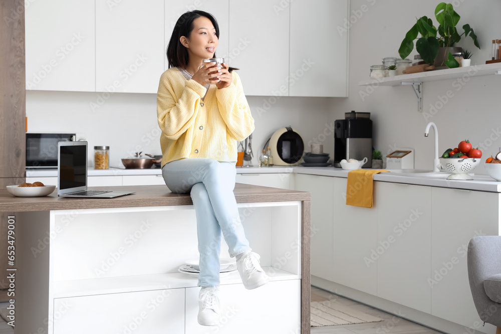 Beautiful young Asian woman with cup of tasty tea sitting at kitchen