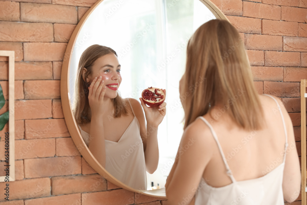 Young woman with pomegranate and under-eye patches near mirror in bathroom