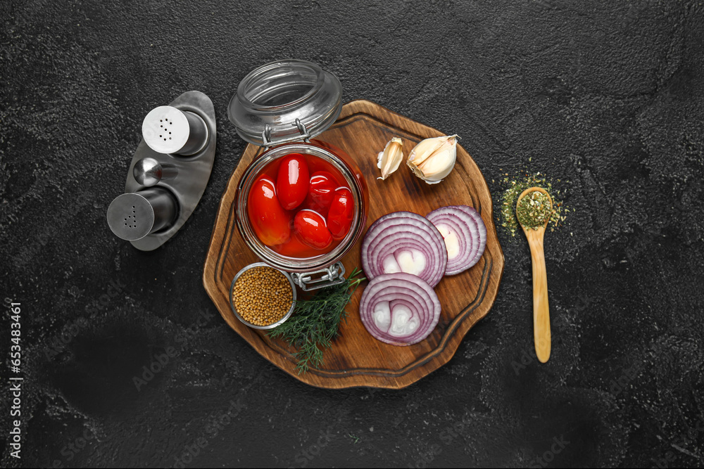 Wooden board with jar of pickled tomatoes and different spices on black background