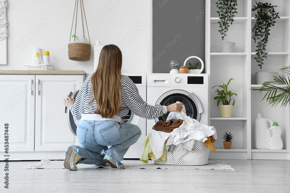 Young woman putting dirty clothes into washing machine in laundry room, back view