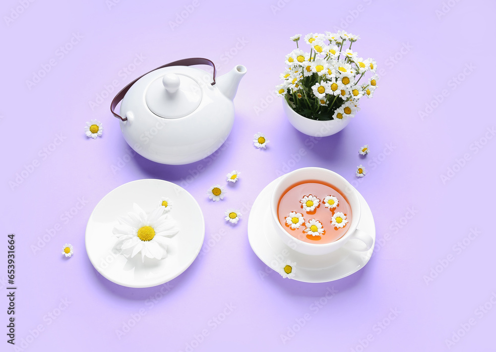 Teapot with cup of natural chamomile tea and flowers on lilac background