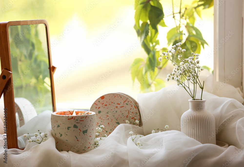 Holder with burning candle, gypsophila flowers and mirror on windowsill in room, closeup