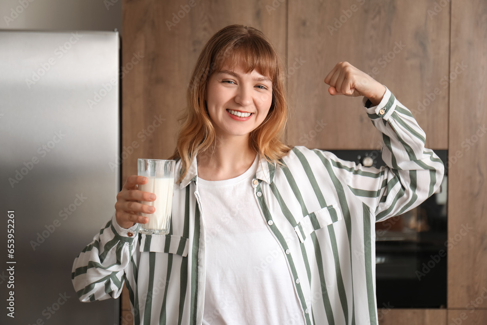 Young woman with glass of milk showing muscles in kitchen