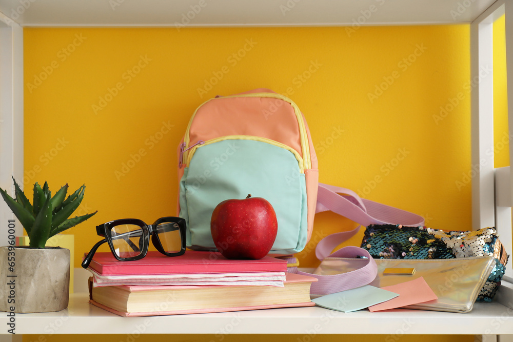 School backpack, stationery, eyeglasses and apple on shelf unit near color wall