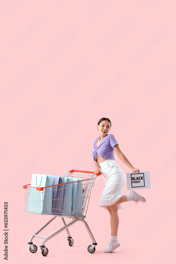 Young Asian woman with shopping bags in cart on pink background. Black Friday sale