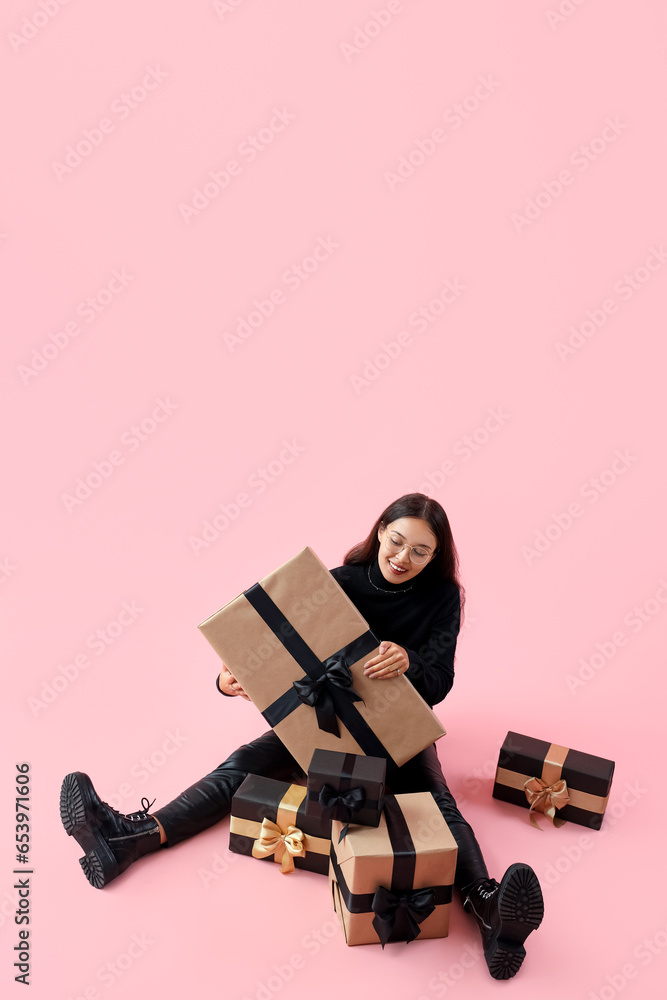 Young Asian woman with gifts sitting on pink background. Black Friday sale