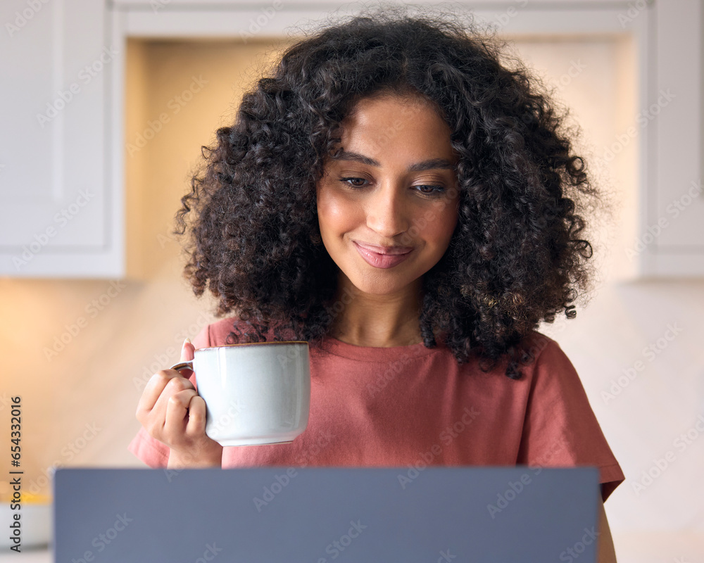 Young Woman In Kitchen At Home Looking At Laptop Holding Hot Drink