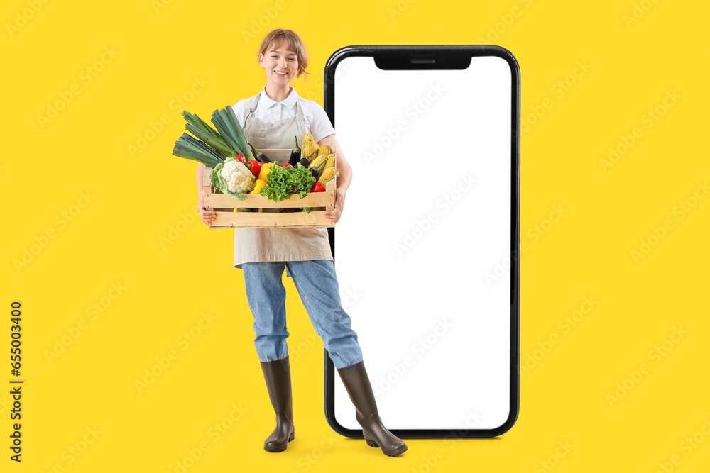 Big smartphone and female farmer with box full of ripe vegetables on yellow background