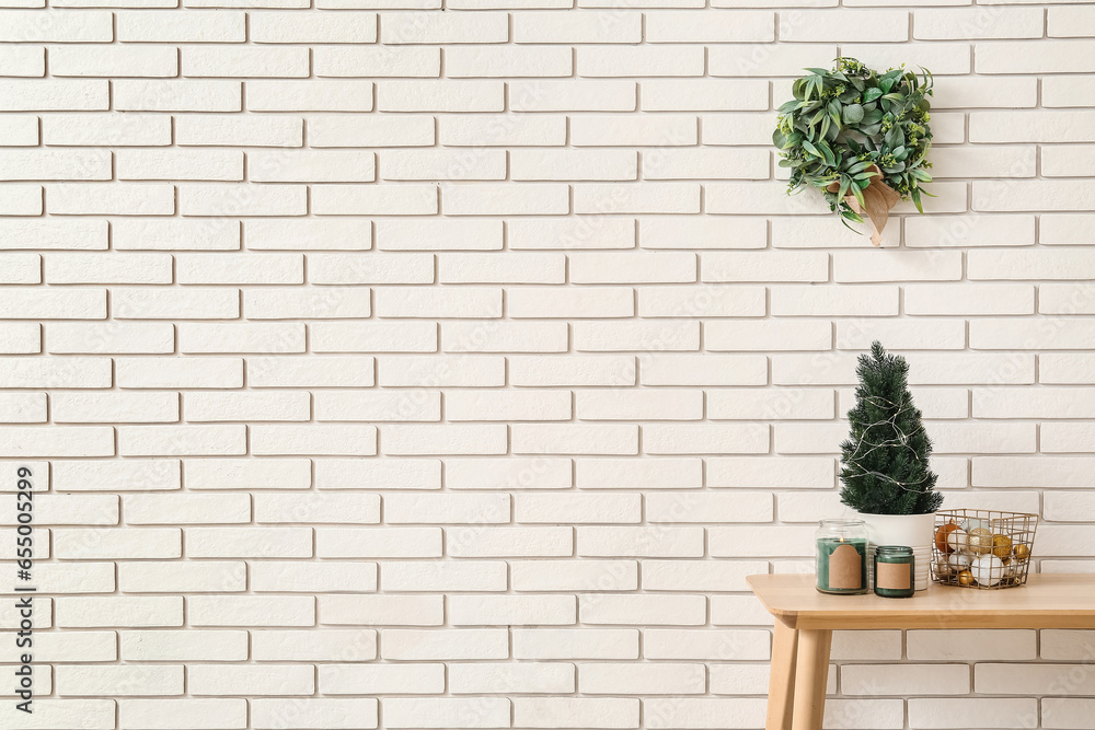 Table with fir tree, candles, Christmas balls and mistletoe wreath on white brick wall