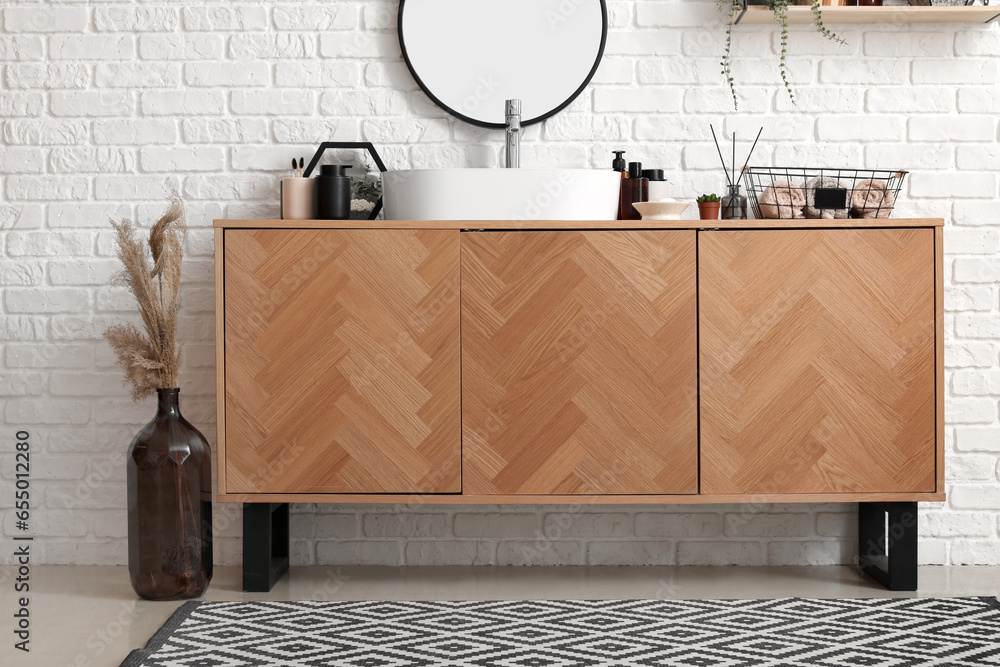 Wooden cabinet with sink bowl and bath accessories near white brick wall