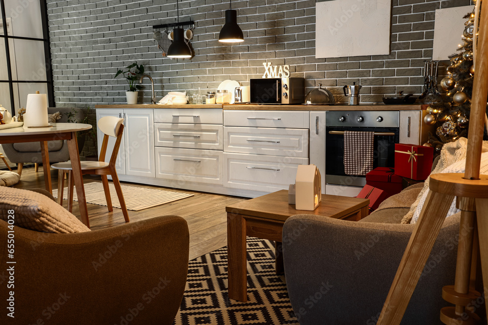 Interior of modern kitchen with Christmas tree, white counters and grey sofa at evening