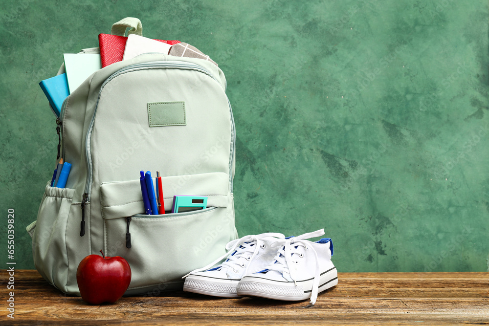 School backpack with notebooks, apple and sneakers on wooden table near green wall