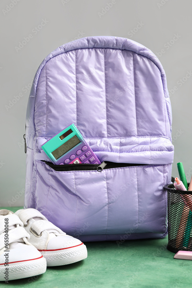 Lilac school backpack with calculator, sneakers and cup of pens on green table near white wall