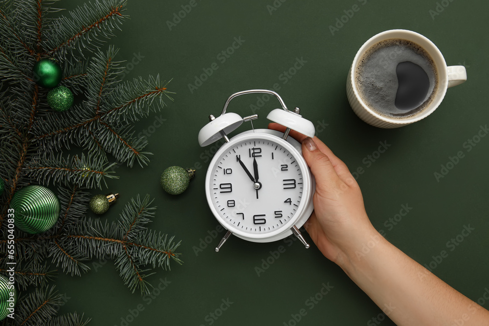 Woman holding alarm clock with Christmas tree branches and cup of coffee on green background