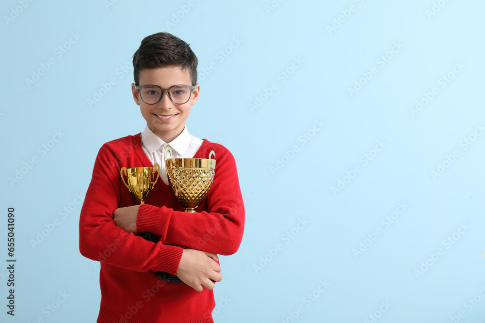 Smart little school boy with prize cups on light blue background