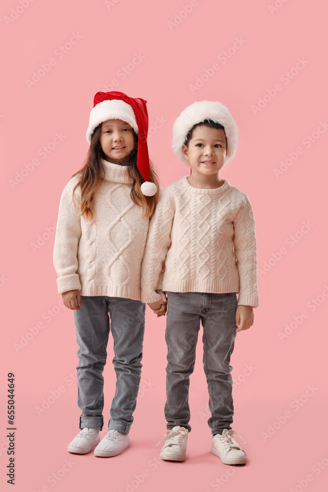 Cute little children in winter sweaters and Santa Claus hats on pink background