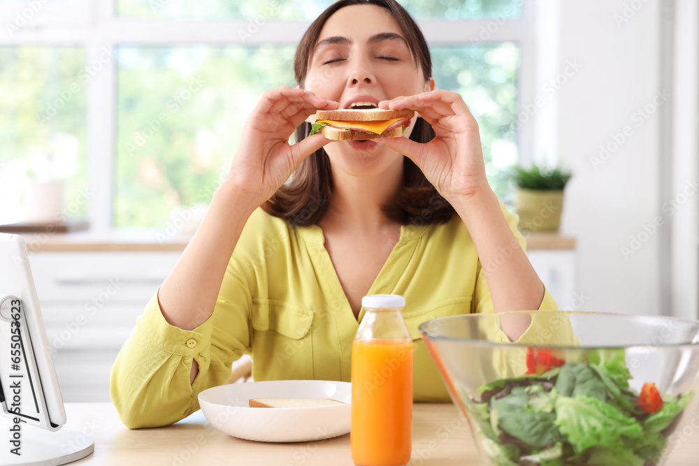 Beautiful young woman eating tasty sandwich in kitchen