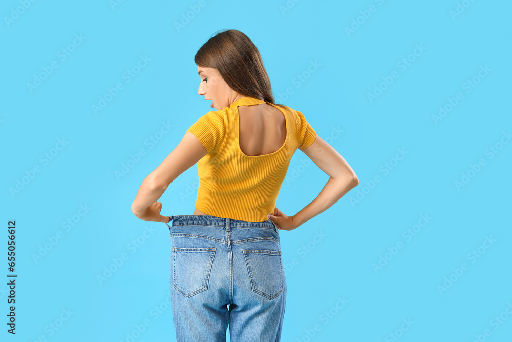 Beautiful young woman in loose jeans on blue background, back view. Weight loss concept