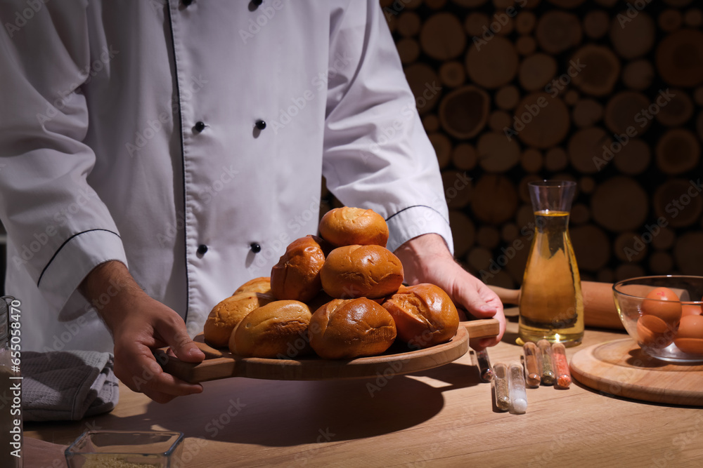 Young chef holding board with fresh buns in bakery, closeup