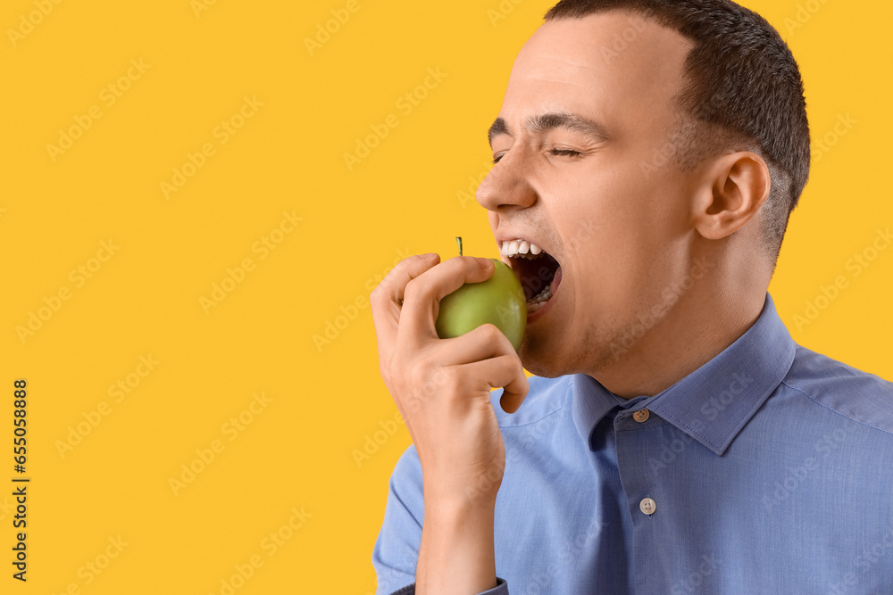 Young man eating tasty apple on yellow background, closeup