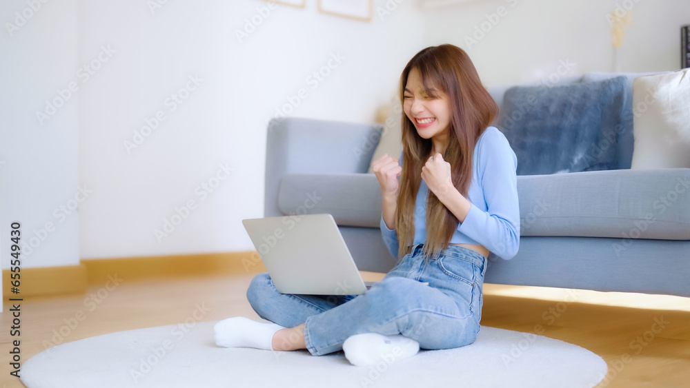 Young asian woman in good spirits working on laptop at home while sitting on the floor close to the couch. Excited female winner celebrating success while glancing at notebook