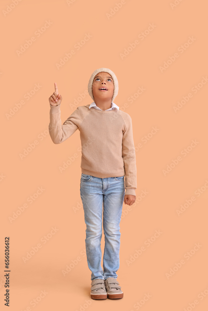 Cute African-American boy in warm winter clothes pointing at something on beige background