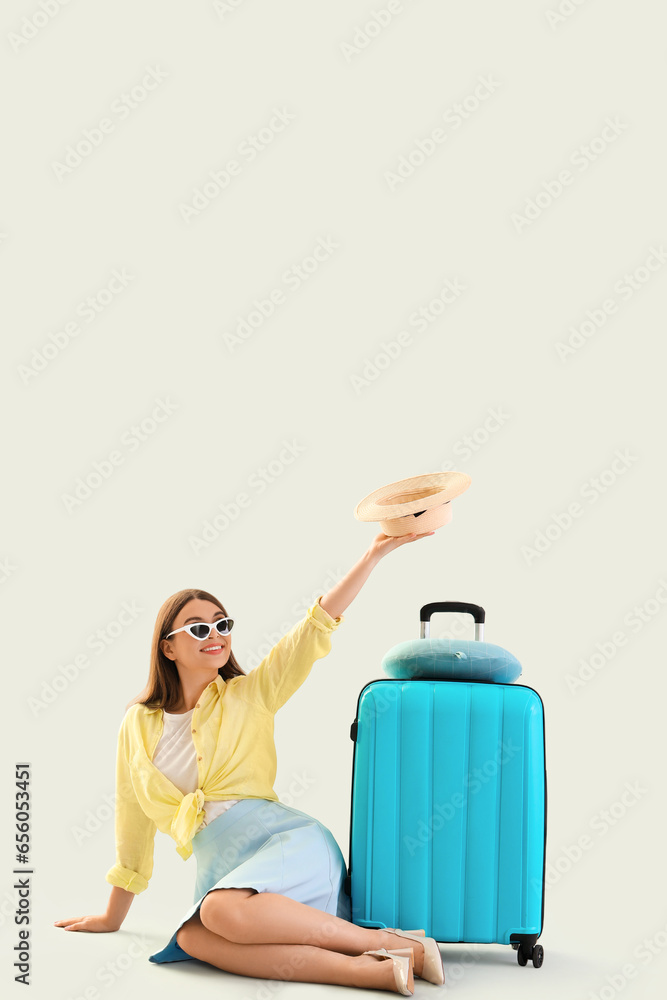 Young woman with summer hat and suitcase sitting on light background