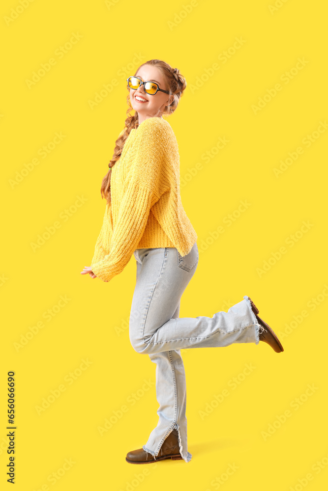 Beautiful happy young woman in warm sweater and eyeglasses on yellow background