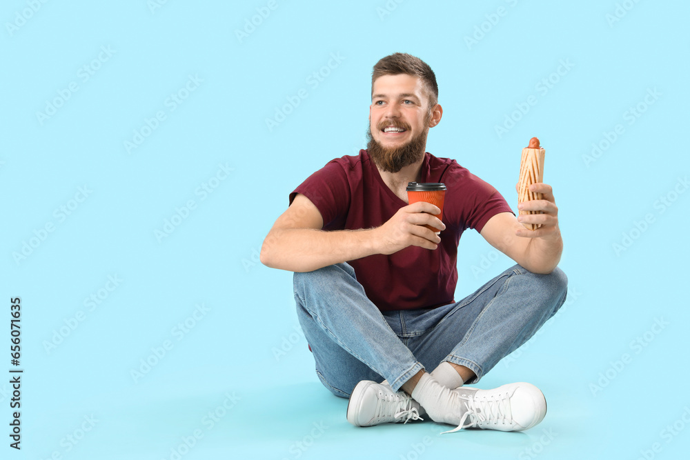 Happy young man with tasty hot dog and cup of coffee sitting on blue background