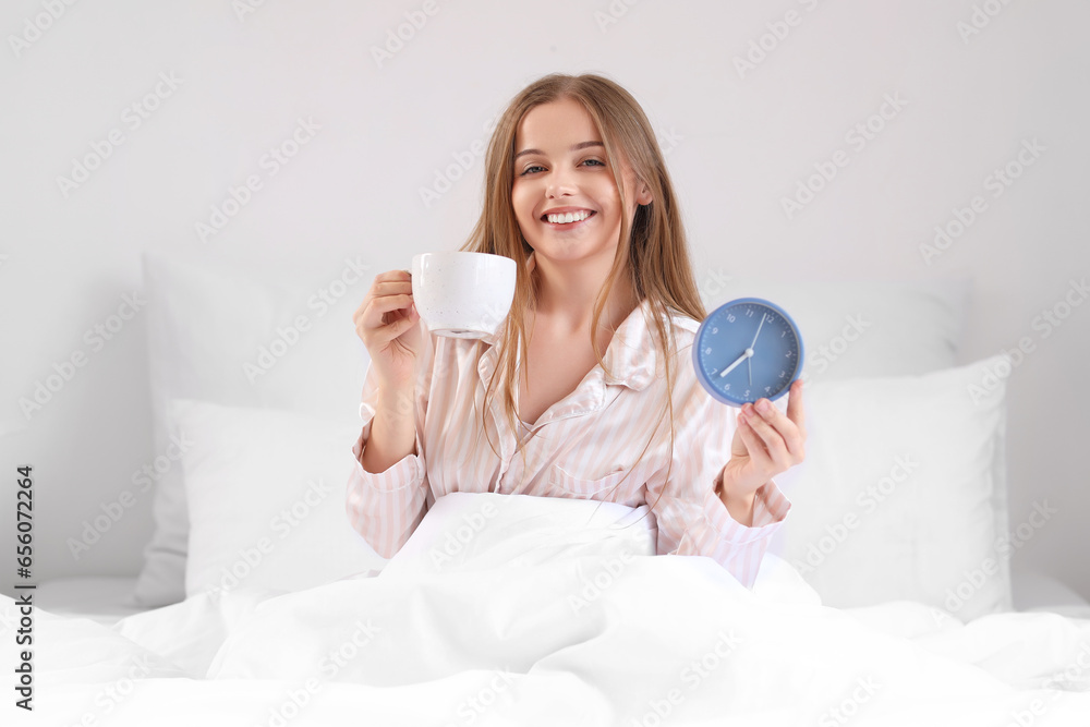 Young woman with cup of coffee and alarm clock in bed at home