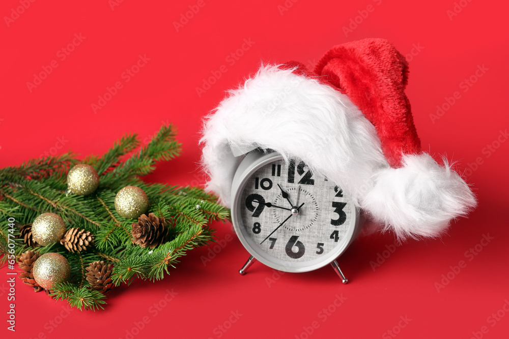 Alarm clock with Santa hat, Christmas tree branches and balls on red background