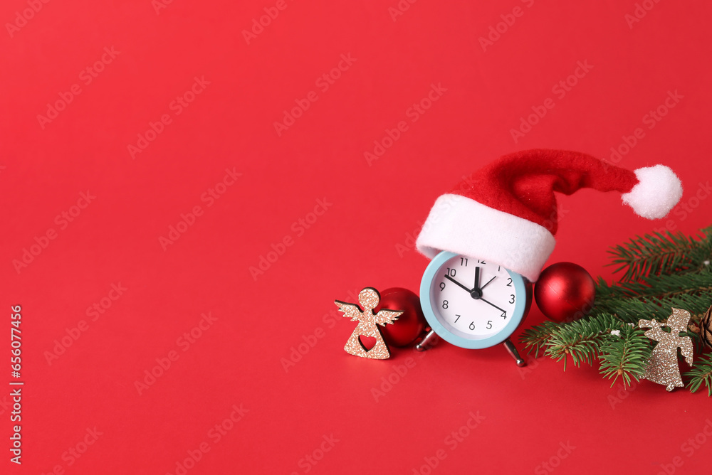 Alarm clock with Santa hat, Christmas tree branches and beautiful decorations on red background