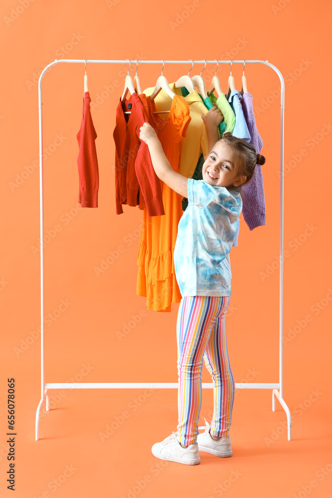 Cute little girl and rack with clothes on orange background