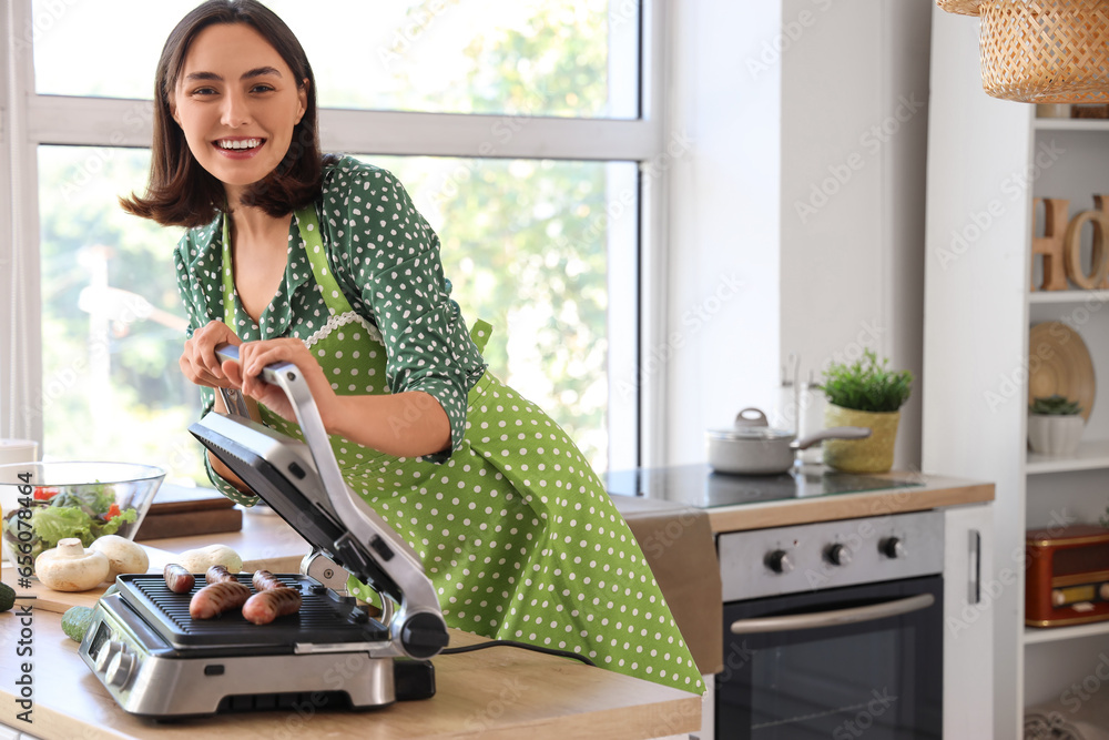 Beautiful young woman cooking delicious sausages on modern electric grill in kitchen