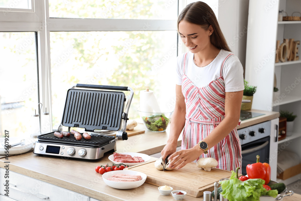 Beautiful young woman cutting mushrooms near modern electric grill with sausages at kitchen