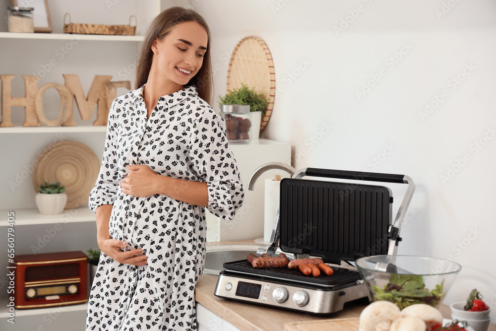 Beautiful young pregnant woman cooking sausages on modern electric grill in kitchen