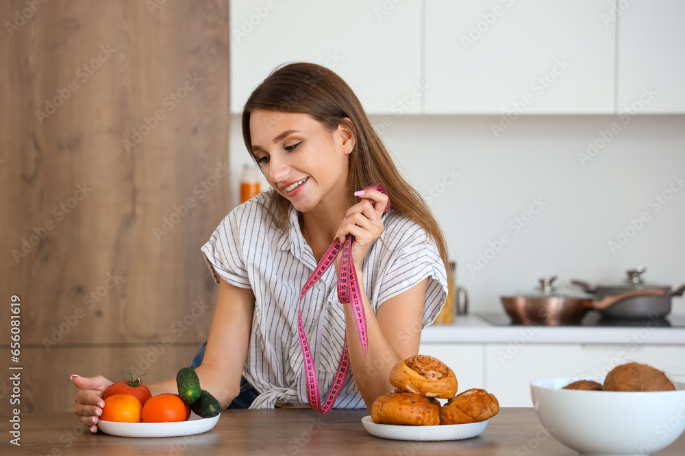 Young woman with measuring tape choosing between healthy and unhealthy food in kitchen. Weight loss concept